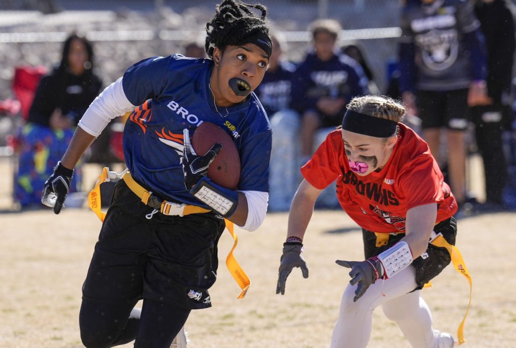 Duas jogadoras de flag football em ação. Uma de azul corre com a bola; a outra de vermelho a persegue em campo.