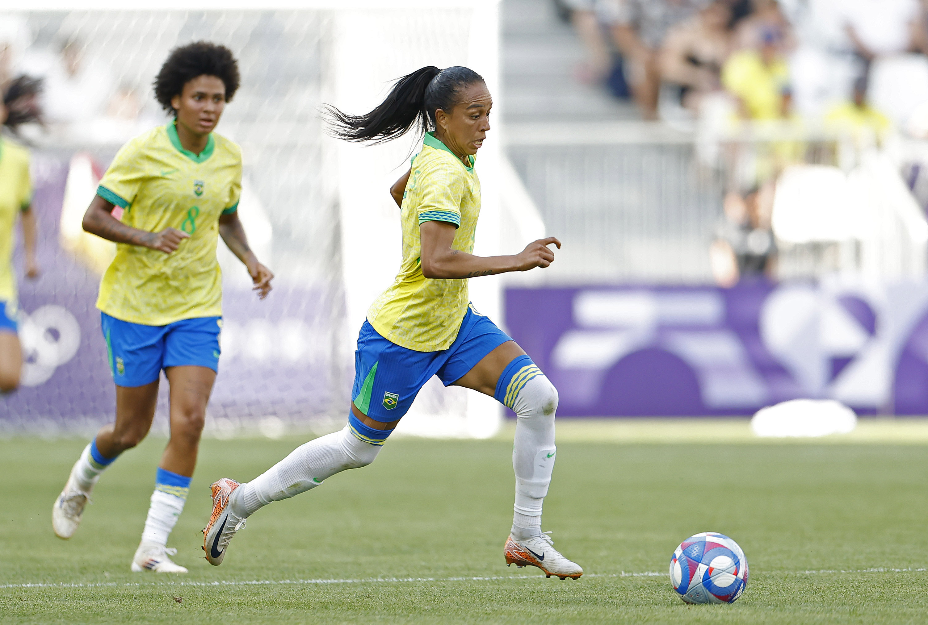 Jogadora de futebol em uniforme amarelo e azul dribla a bola em campo, outra jogadora ao fundo.