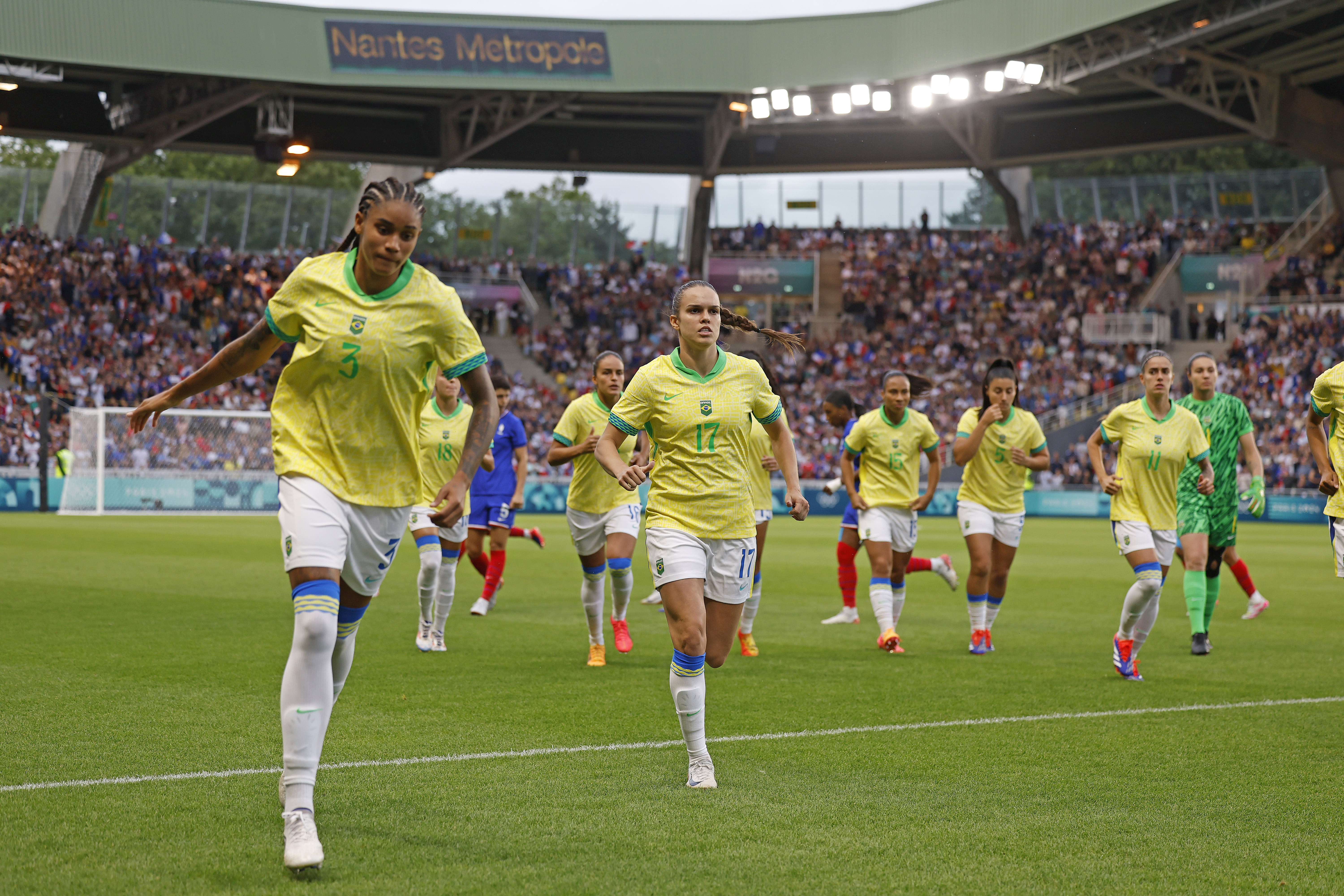Jogadoras de futebol em uniformes amarelos correm no gramado de um estádio. Ao fundo, torcedores e o letreiro "Nantes Metropole".