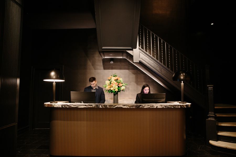 Dois recepcionistas em um balcão com computadores e flores. Uma escada escura ao fundo.