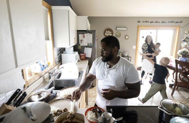 Homem serve refeição em cozinha movimentada, com louça suja. Ao fundo, mulher segura bebê e criança brinca.