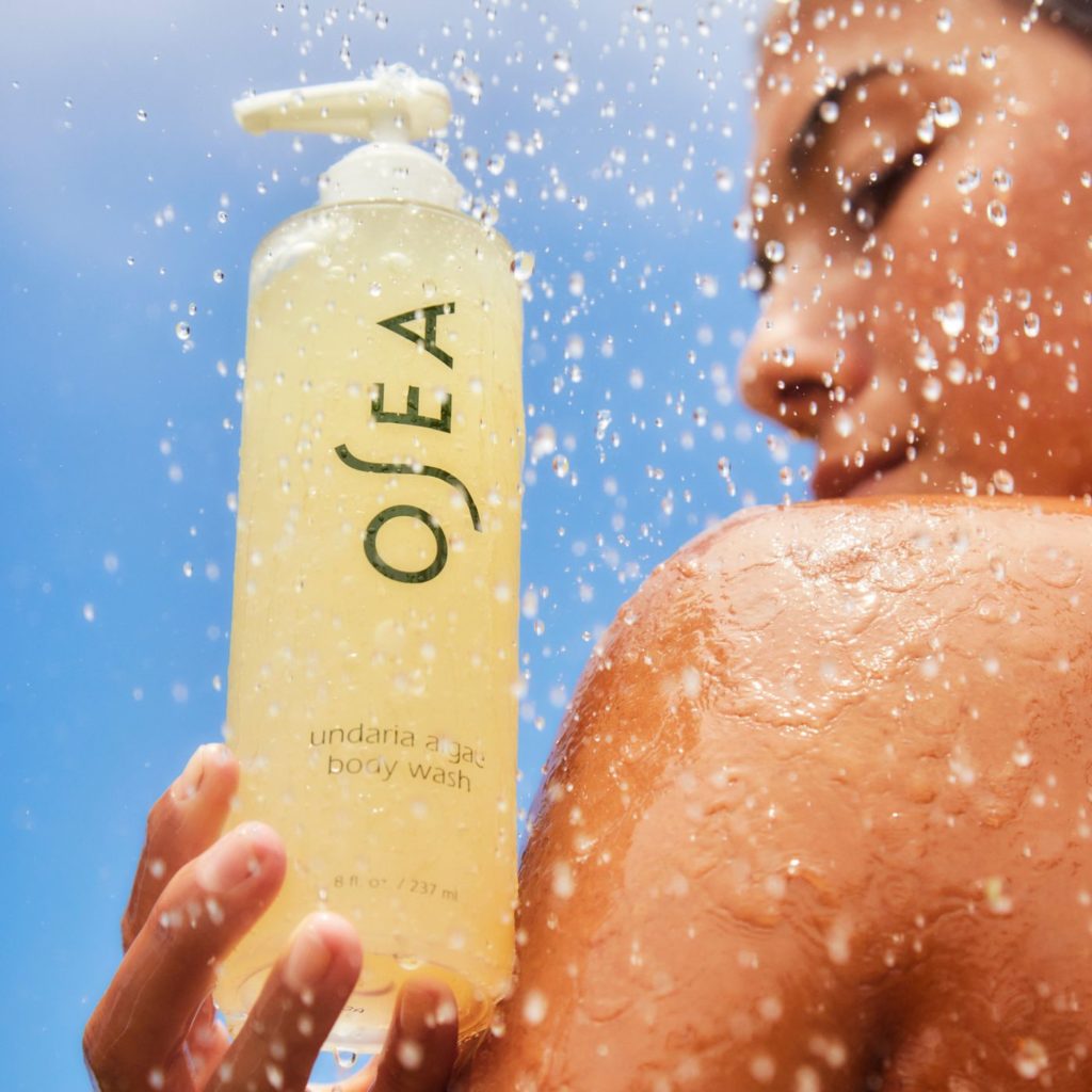 Mulher se banha sob água, segurando uma garrafa de OSEA body wash. Gotas e céu azul ao fundo.