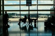 Pessoas silhuetadas esperam em aeroporto, sentadas perto de janelas com aviões na pista ao fundo.
