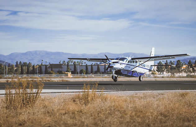 Avião branco e azul decolando. Pista e grama seca em primeiro plano, montanhas ao fundo.