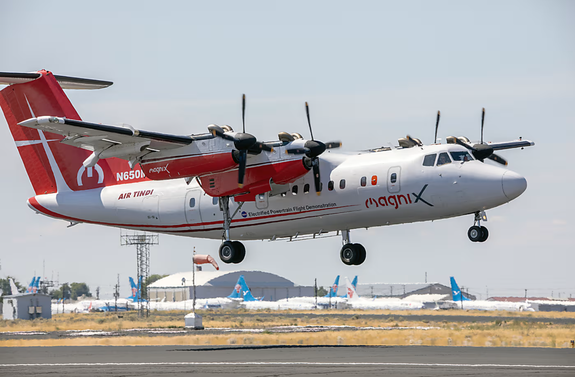 Avião elétrico branco e vermelho da magniX pousando. Aeronave com hélices e trem de pouso estendido, próxima ao solo.