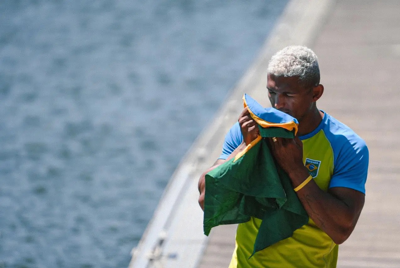Atleta de cabelo branco beija bandeira do Brasil. Ao fundo, água e cais.