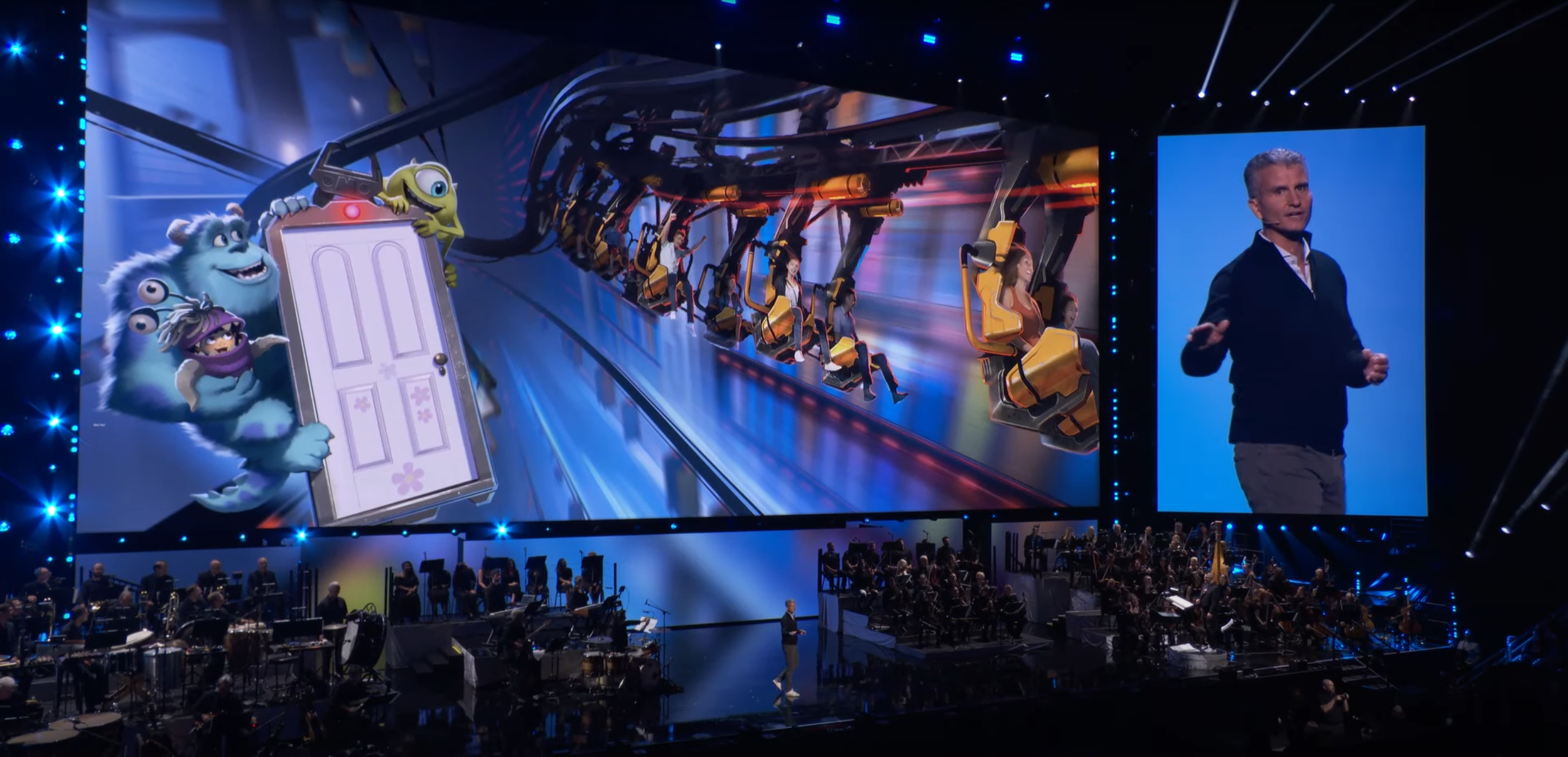 Tela grande exibe Monstros S.A. e uma atração futurista; palco com orquestra e apresentador.