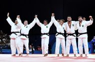 Seis judocas brasileiros comemoram vitória, erguendo os braços unidos no tatame.