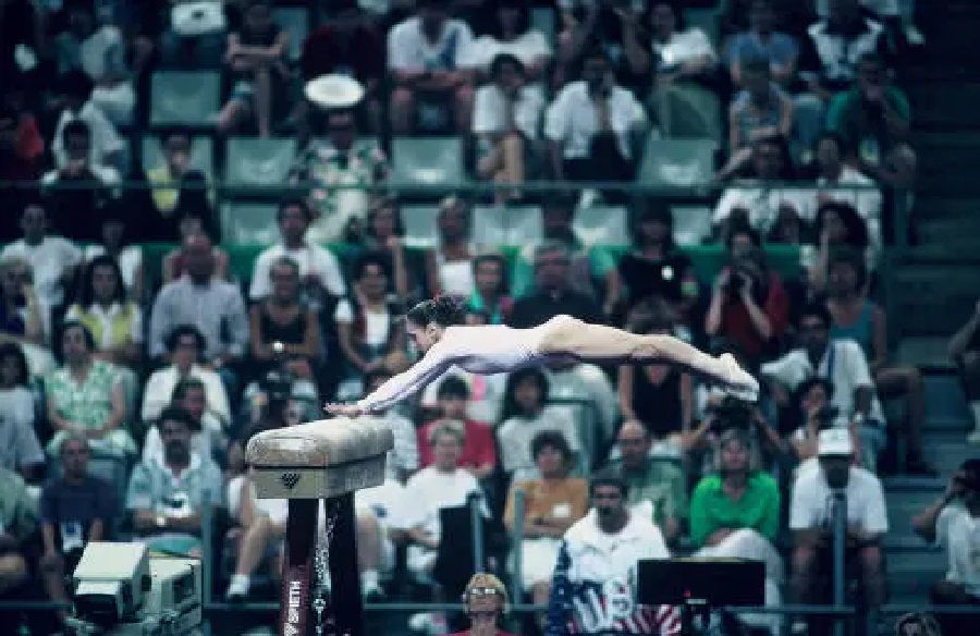 Ginasta feminina em salto na mesa de salto, com público ao fundo.