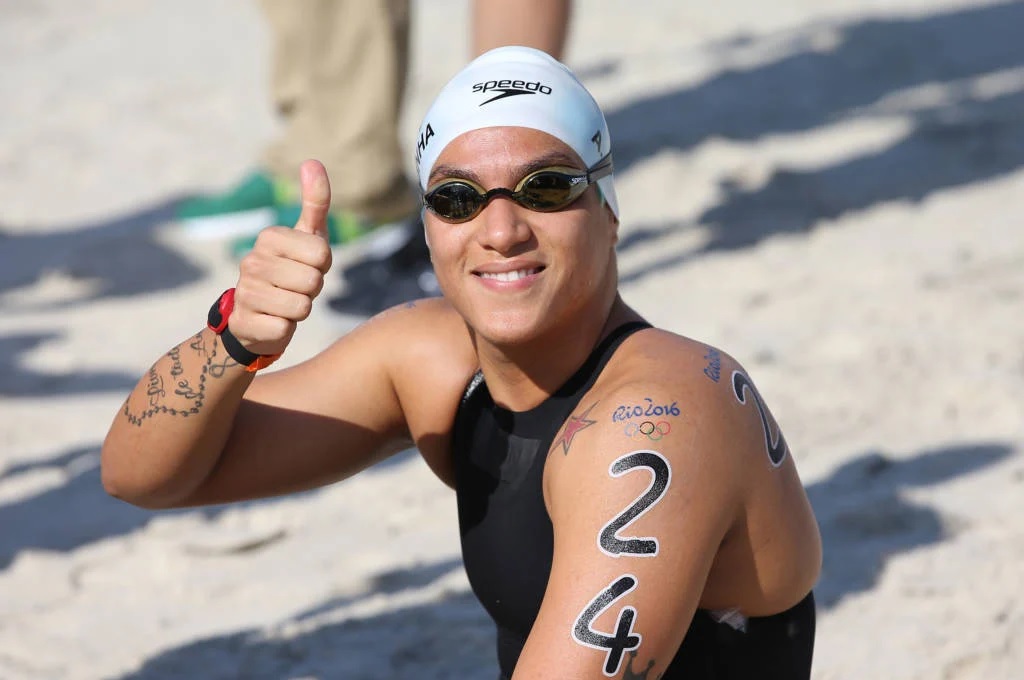 Triatleta sorridente com touca e óculos de natação, mostrando polegar para cima. Número 24 no braço.