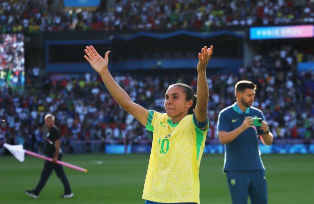 Jogadora de futebol brasileira, de braços erguidos no gramado, saúda a torcida em estádio lotado.