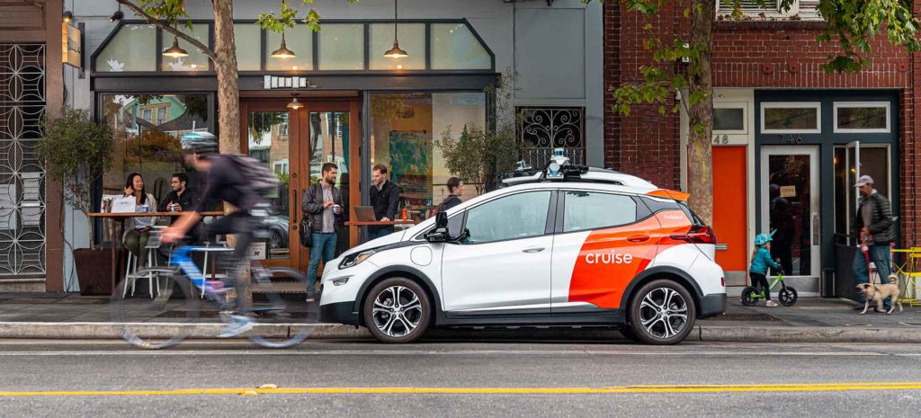 Carro autônomo branco e laranja em rua. Pessoas próximas em movimento.