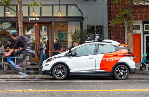 Carro autônomo branco e laranja em rua. Pessoas próximas em movimento.