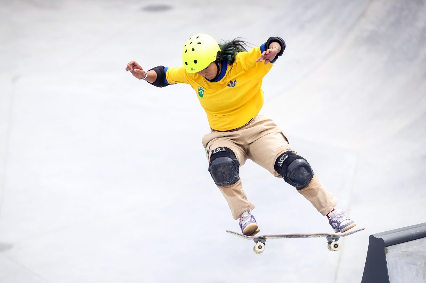 Uma skater brasileira com capacete amarelo e camisa do Brasil executa manobra no ar em skatepark.