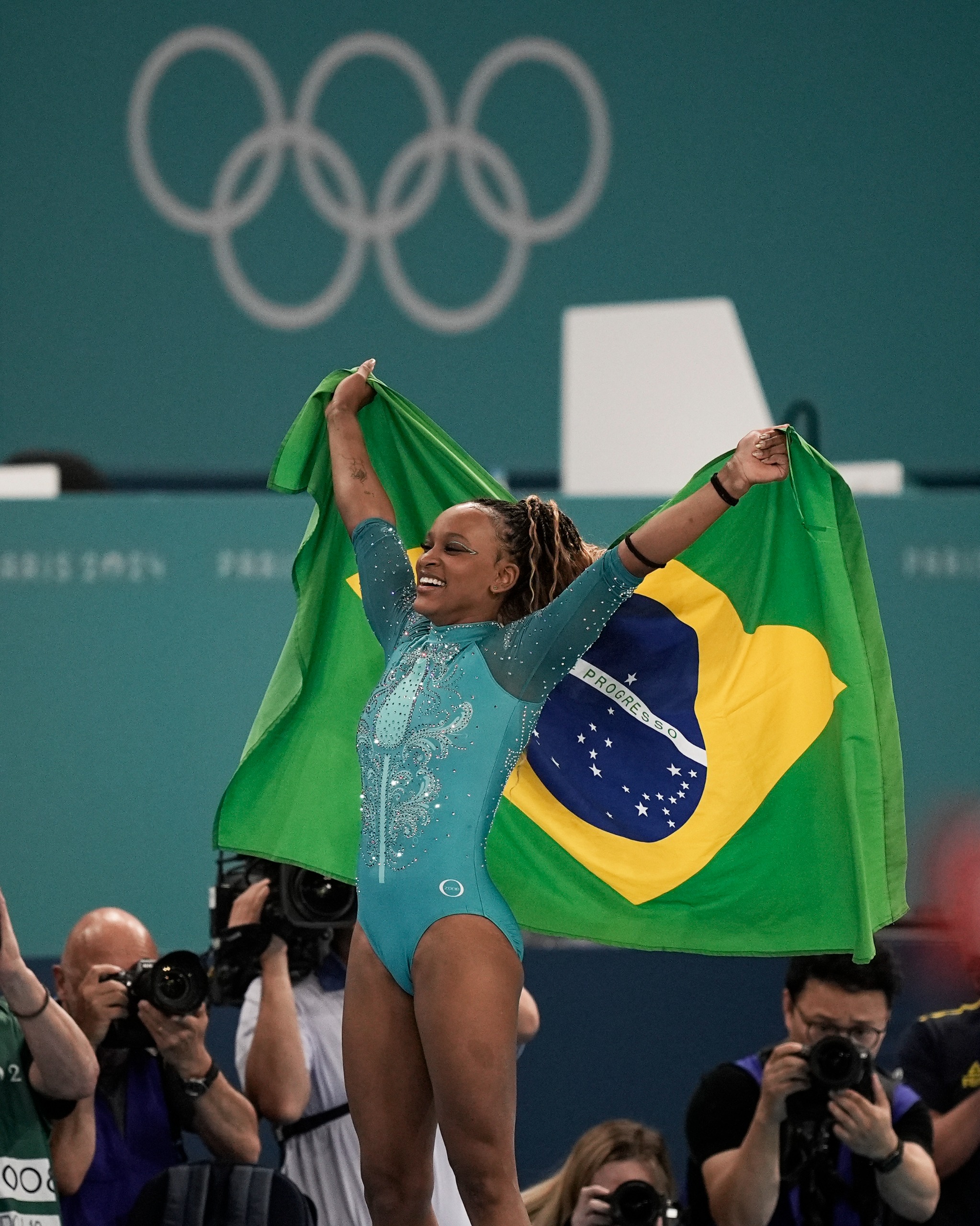 Ginasta feminina sorrindo, segurando a bandeira do Brasil acima da cabeça, com anéis olímpicos ao fundo.