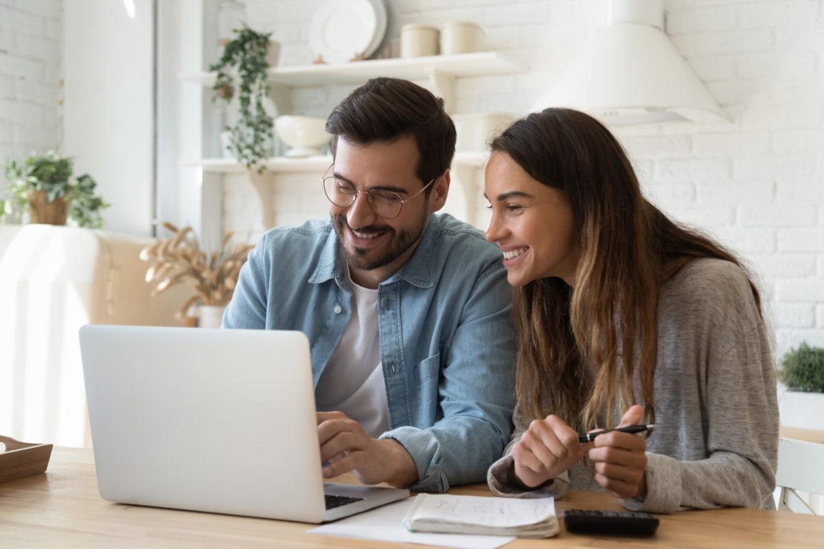 casal de homem e mulher sorrindo em frente a um notebook com tela aberta. na mesa há um caderno e uma calculadora