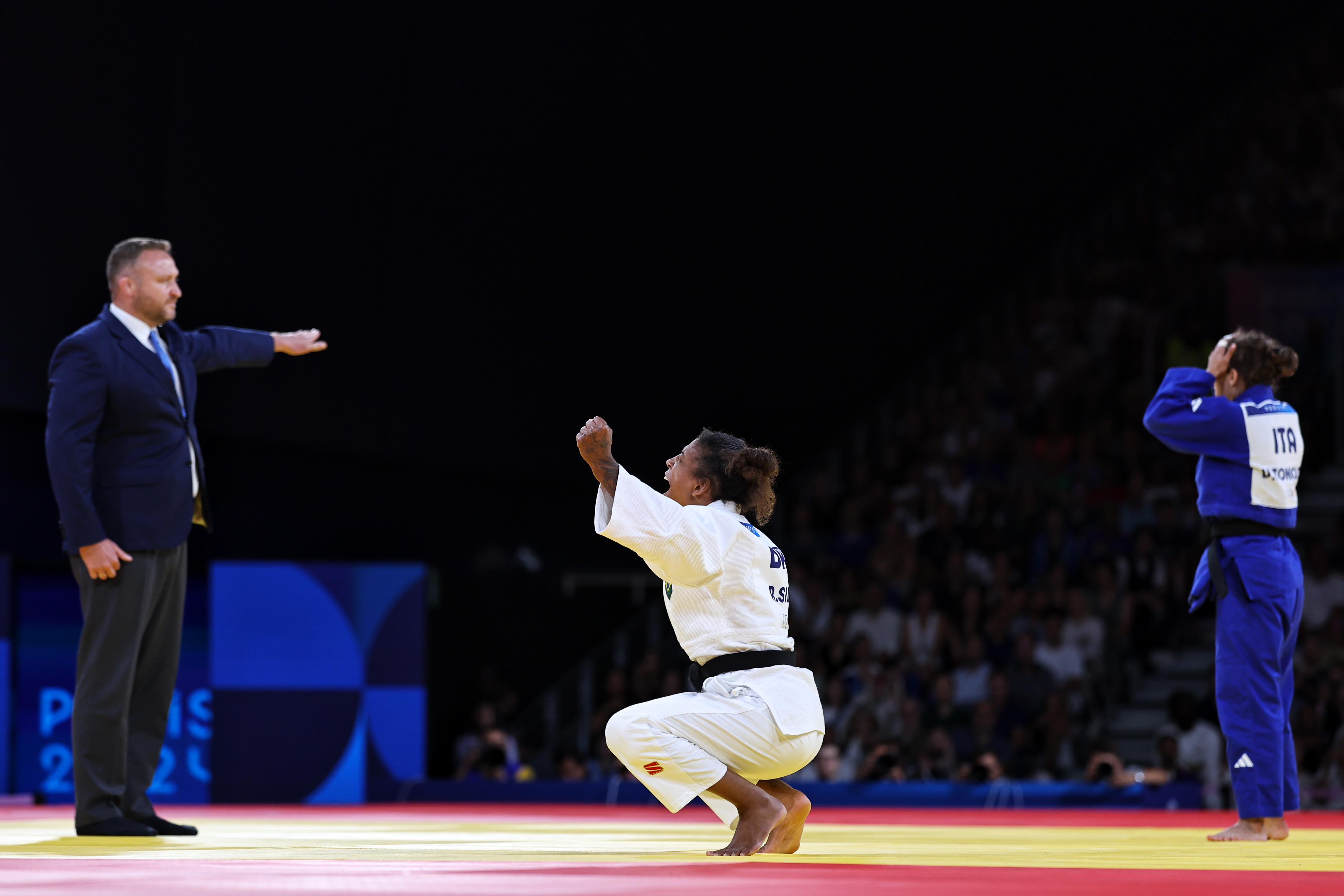 Em luta de judô, atleta de branco celebra agachada, outra de azul lamenta em pé. Árbitro aponta.