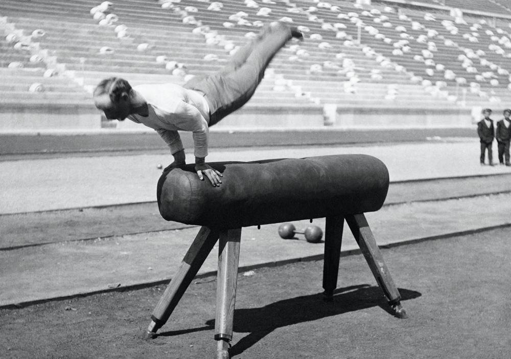 Ginasta faz parada de mãos em cavalo de ginástica. Fundo com arquibancadas de estádio.