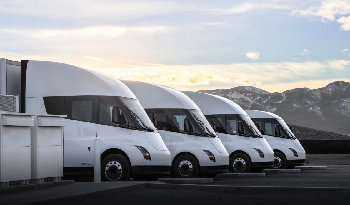 Quatro caminhões Tesla Semi brancos estacionados em fila, ao pôr do sol, com montanhas ao fundo.