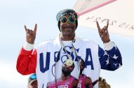 Snoop Dogg sorrindo, óculos olímpicos, jaqueta EUA com atletas, fazendo sinal de rock.