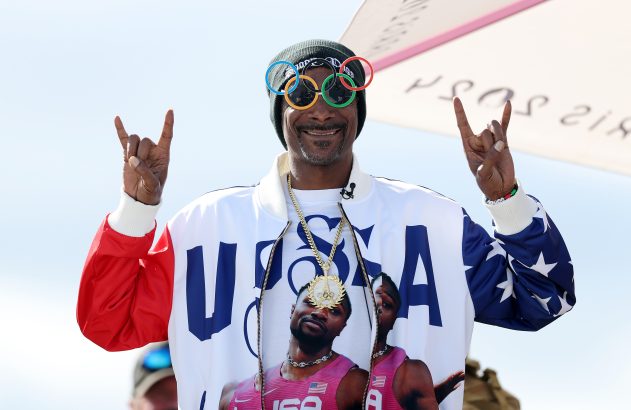 Snoop Dogg sorrindo, óculos olímpicos, jaqueta EUA com atletas, fazendo sinal de rock.