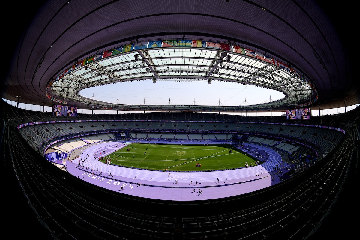 Vista panorâmica do estádio Paris 2024. Pista de atletismo roxa, campo verde com anéis olímpicos. Pessoas na pista.