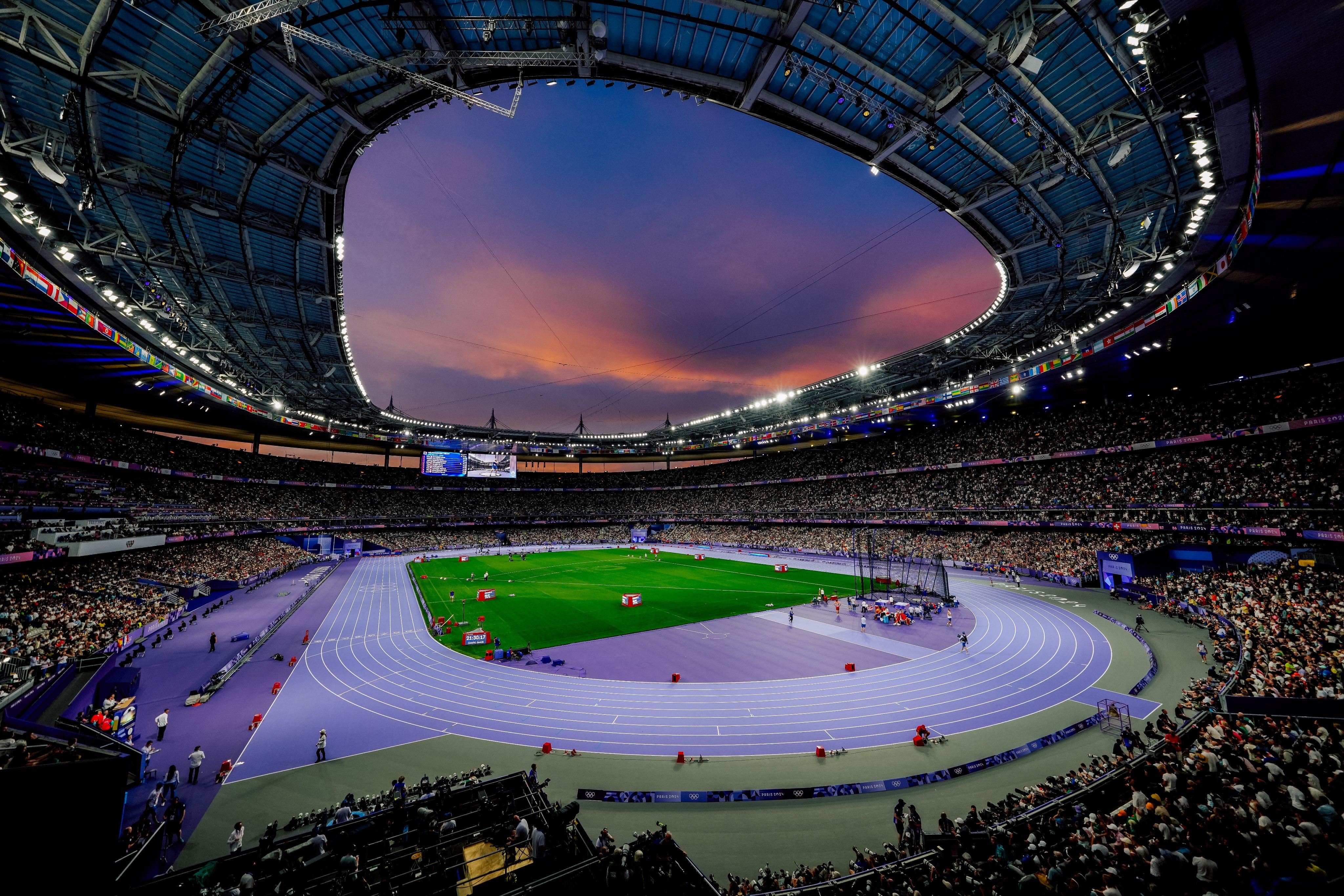 Estádio de atletismo lotado ao pôr do sol, com pista roxa e gramado verde sob céu colorido.