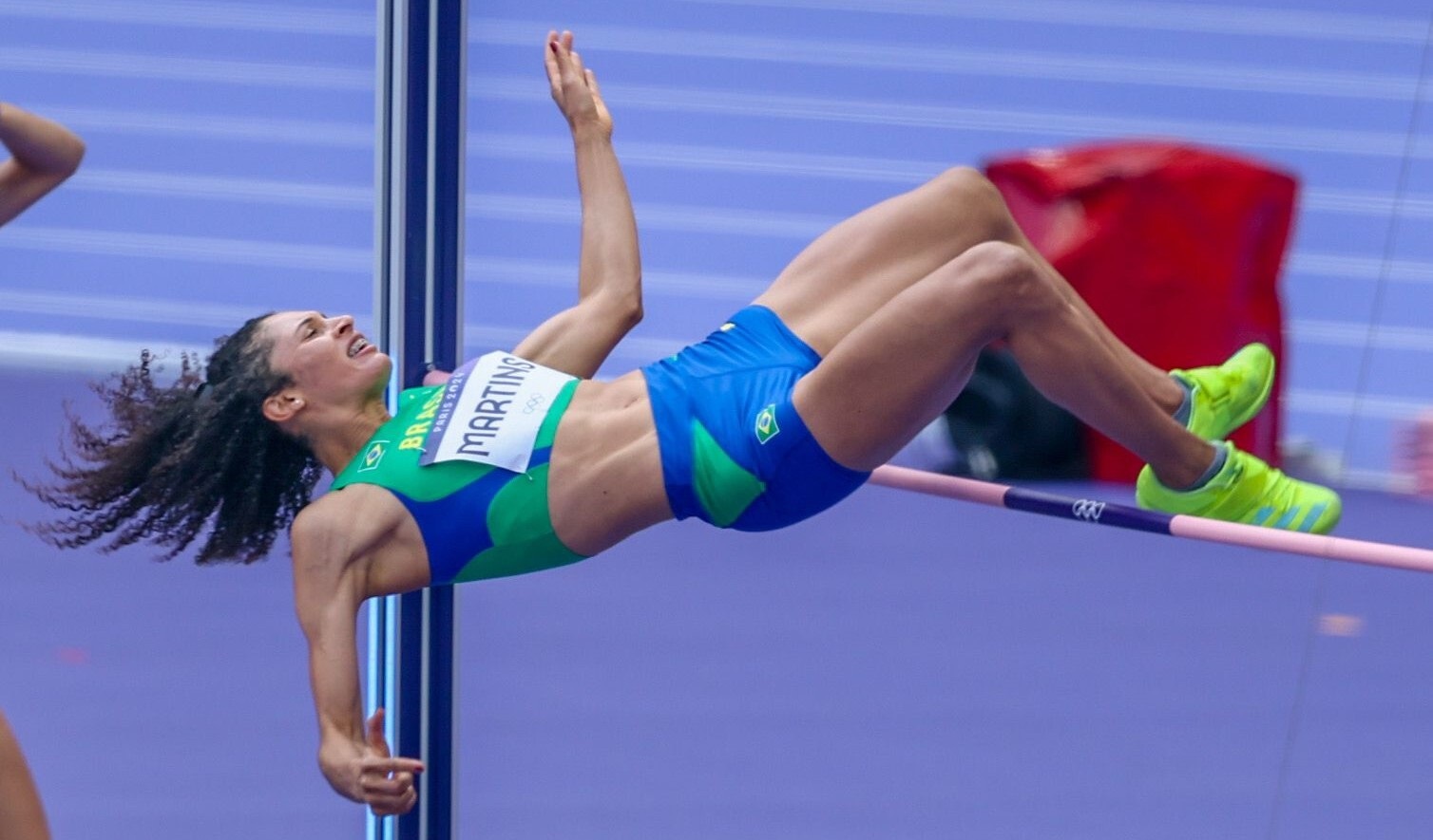 Atleta feminina de salto em altura em uniforme verde e azul, saltando de costas sobre a barra.