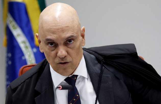Homem careca de terno e toga preta, olhar sério, em sessão com microfone e bandeira do Brasil.