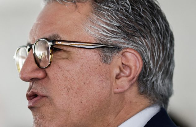 Homem de perfil, óculos, cabelo grisalho, boca aberta como se falasse. Veste terno. Fundo claro desfocado.