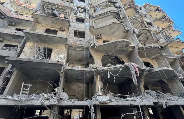 Prédio de múltiplos andares severamente destruído, com paredes ausentes expondo o interior e escombros.
