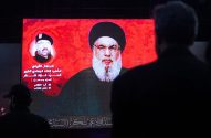 Pessoas observam telão vermelho exibindo retratos de um clérigo e outro líder com texto árabe de homenagem.