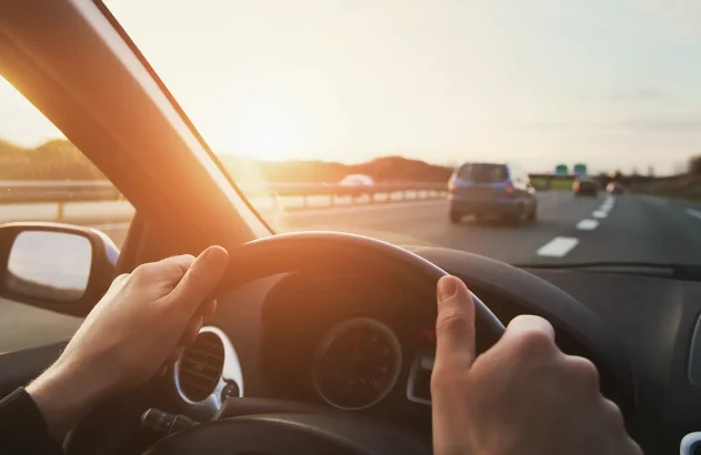 Close do painel de um carro. Em primeiro plano, vê-se o close de duas mãos ao volante e ao fundo uma autoestrada com carros circulando