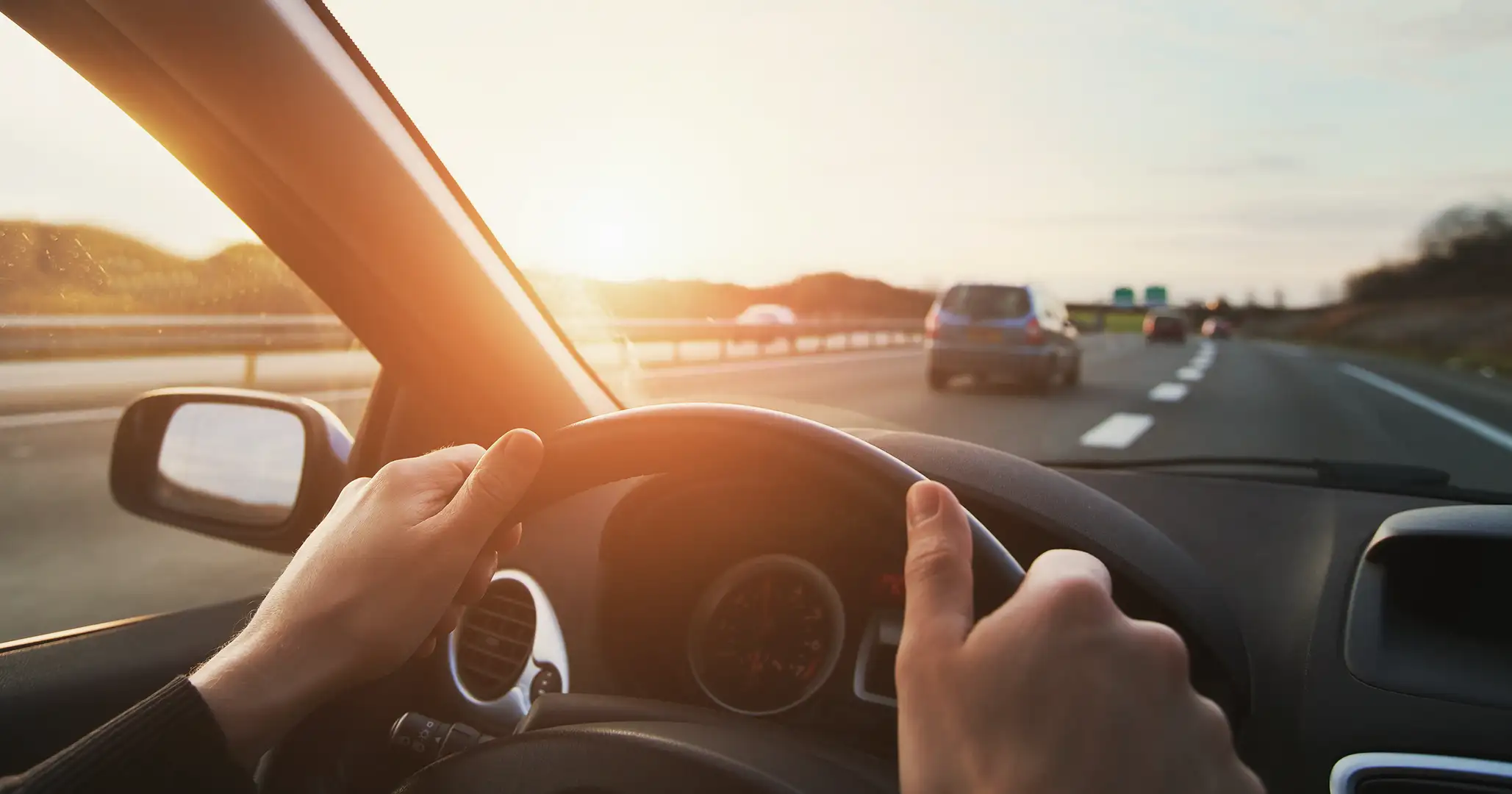 Close do painel de um carro. Em primeiro plano, vê-se o close de duas mãos ao volante e ao fundo uma autoestrada com carros circulando