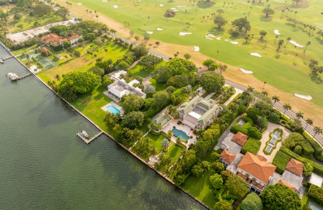 Vista aérea de mansões luxuosas à beira-mar com piscinas e docas, adjacente a um campo de golfe verdejante.