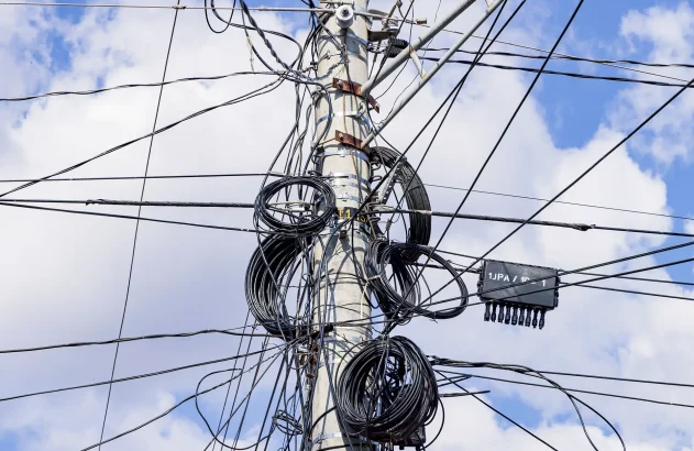 Emaranhado de cabos de energia elétrica presos no alto de um poste de concreto com um céu azul com nuvens ao fundo