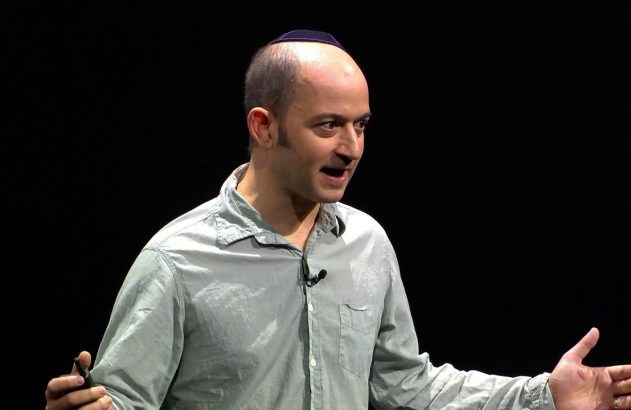 Homem careca de camisa clara, com microfone, discursando em palco escuro.