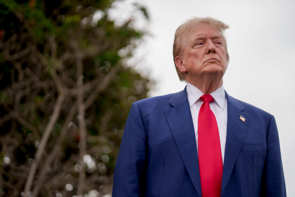 Donald Trump de terno azul e gravata vermelha olhando para cima, ao ar livre.