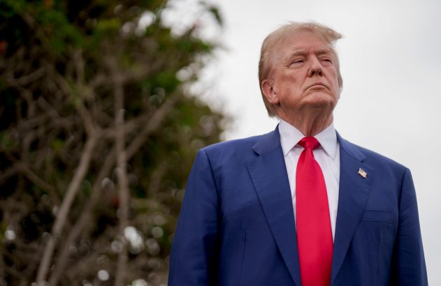 Donald Trump de terno azul e gravata vermelha olhando para cima, ao ar livre.