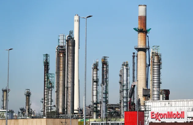 Uma visão geral da refinaria Exxon Mobil no Porto de Roterdã. Na imagem se vê a fachada da edificação, feita de metal e alvenaria, com o letreiro da empresa à direita, contra um céu azul.
