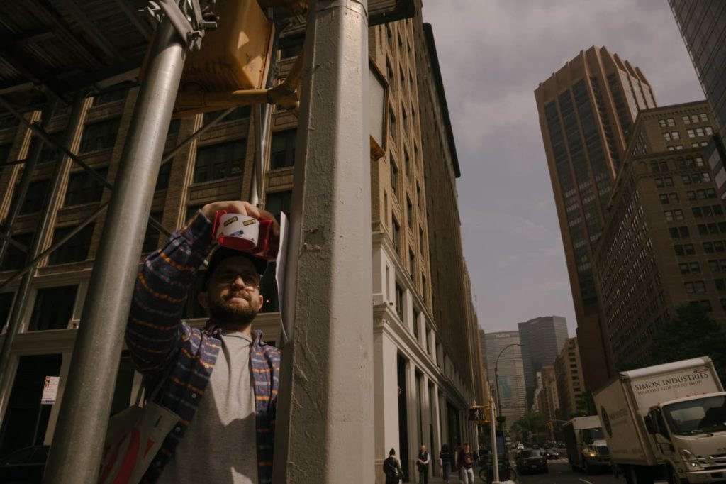 Homem cola um papel em um poste, usando um dispensador de fita. Cena urbana com prédios altos ao fundo.