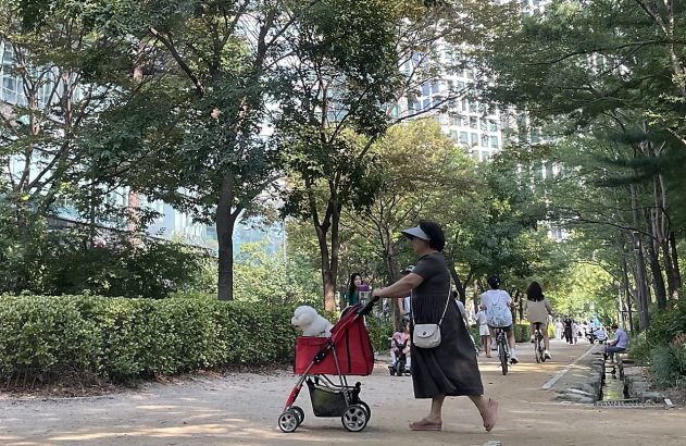 Mulher empurra carrinho vermelho em caminho com árvores sob sol.