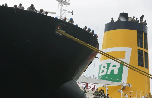 Recorte de um navio petroleiro com trabalhadores na borda do convés, observando o porto. Em segundo plano, uma chaminé amarela com o logo da BR distribuidora.