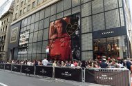 Grande fila em frente à loja Coach na 685 Fifth Avenue, com banner de Selena Gomez e barreiras Coach.