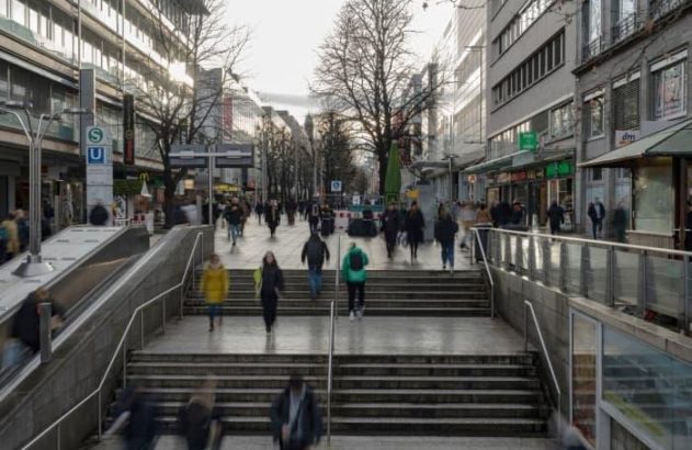 Rua movimentada com pessoas subindo/descendo escadas e escadas rolantes para o metrô, rodeada por lojas e prédios.