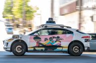Carro autônomo Waymo branco, modelo Jaguar I-Pace, com arte lateral colorida, em movimento numa rua urbana.