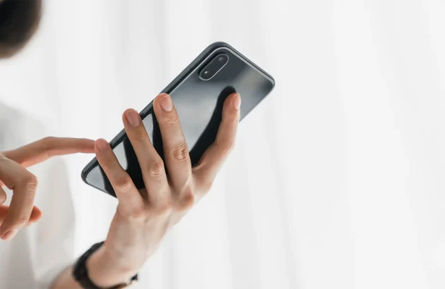 Uma pessoa interage com um smartphone moderno, segurando-o com as duas mãos sobre um fundo branco iluminado.
