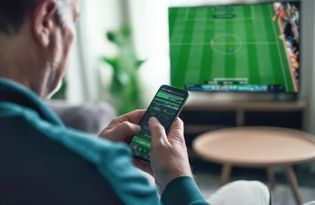 Pessoa de costas e de perfil segurando um celular com aplicativo de apostas esportivas, enquanto uma TV ao fundo exibe uma partida de futebol ao vivo, em um ambiente interno aconchegante com mobiliário moderno.