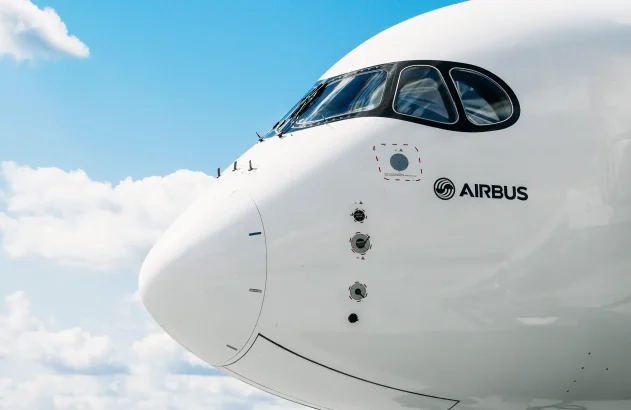 Parte frontal de uma aeronave da Airbus contra um céu azul com algumas nuvens esparsas, mostrando o nariz do avião, a cabine de comando com janelas curvas e o logotipo da Airbus na fuselagem branca.