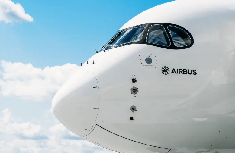 Parte frontal de uma aeronave da Airbus contra um céu azul com algumas nuvens esparsas, mostrando o nariz do avião, a cabine de comando com janelas curvas e o logotipo da Airbus na fuselagem branca.